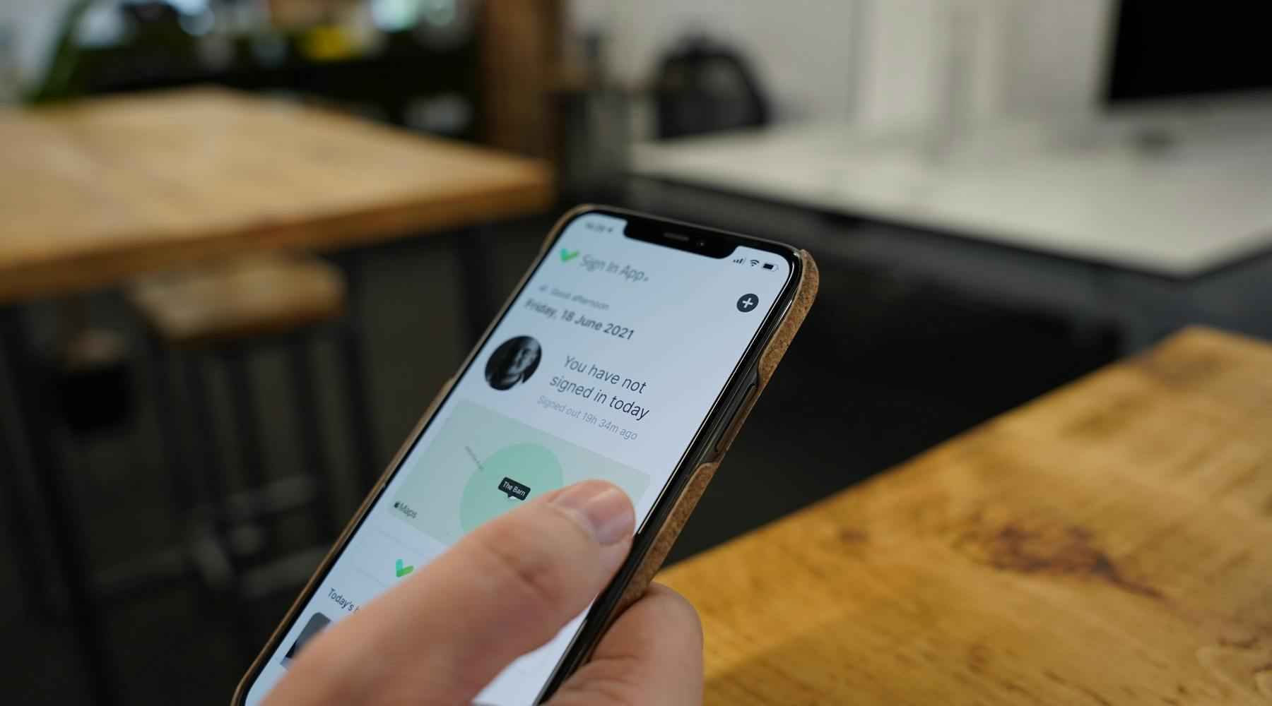 A person holds a smartphone displaying the Sign In App Companion App screen reading "You have not signed in today," against a blurred background of wooden tables in an indoor setting.