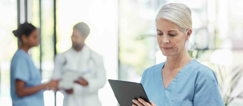 Healthcare worker in scrubs reviewing a tablet in a bright clinic hallway, reflecting the digital, auditable record-keeping HIPAA-compliant visitor management is built to support.