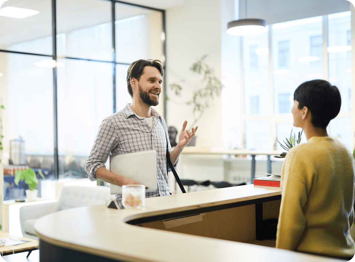 A man holds a laptop, engaging in conversation with a colleague while signing in at a curved reception desk in a bright office setting with large windows and indoor plants.