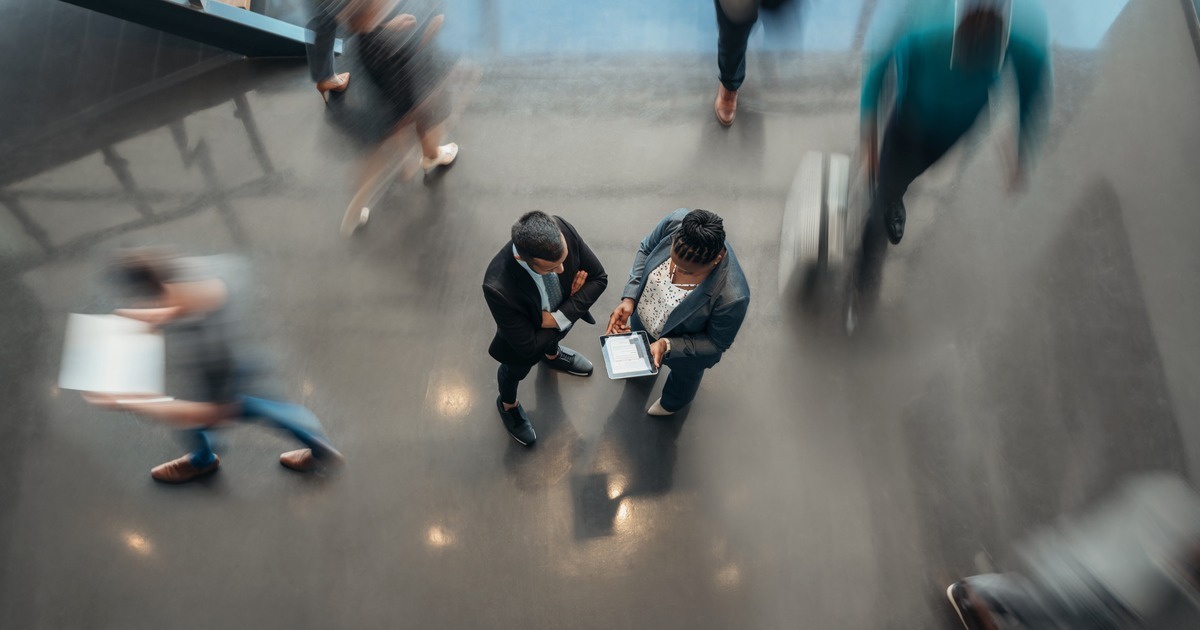 Overhead view of two professionals reviewing a tablet in a busy corporate lobby with people moving around them