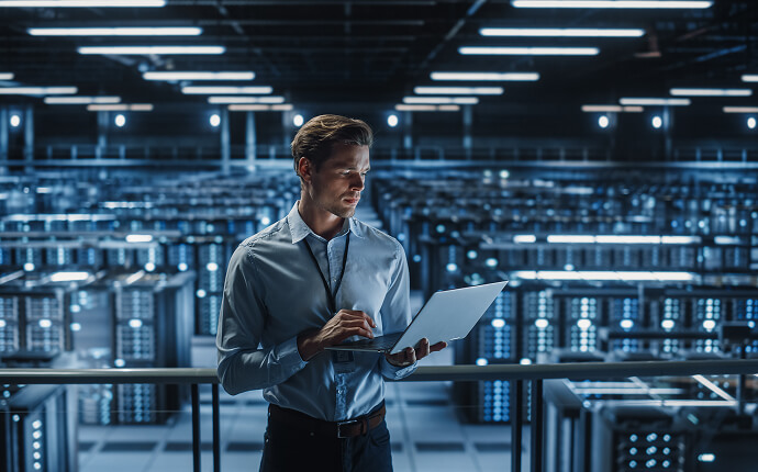 A person stands holding a laptop, in a highly regulated environment with lots of server racks and IT equipment in the background