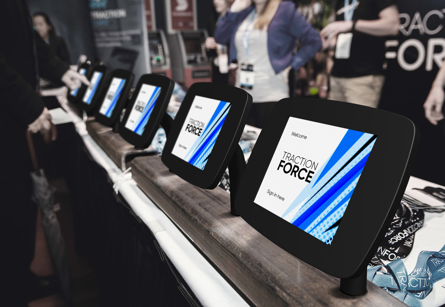 An extrance foyer of an event is shown, with a row of multiple sign in kioks ready for attendees to sign in. Behind some staff can be shown, ready to distribute the guests' badges after signing in
